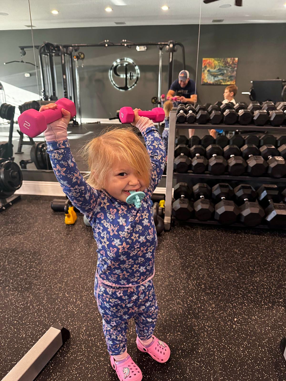 Toddler lifting two small pink dumbbells next to the dumbbell rack